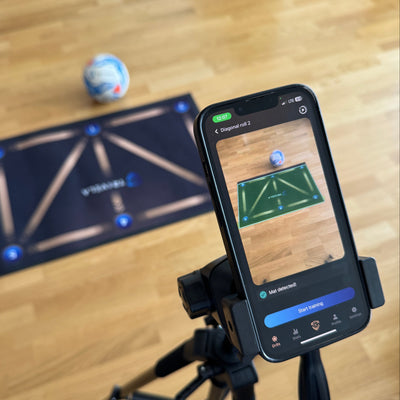 Phone on a tripod with the TRIVELA sports app interface on a wooden floor with a ball and mat.